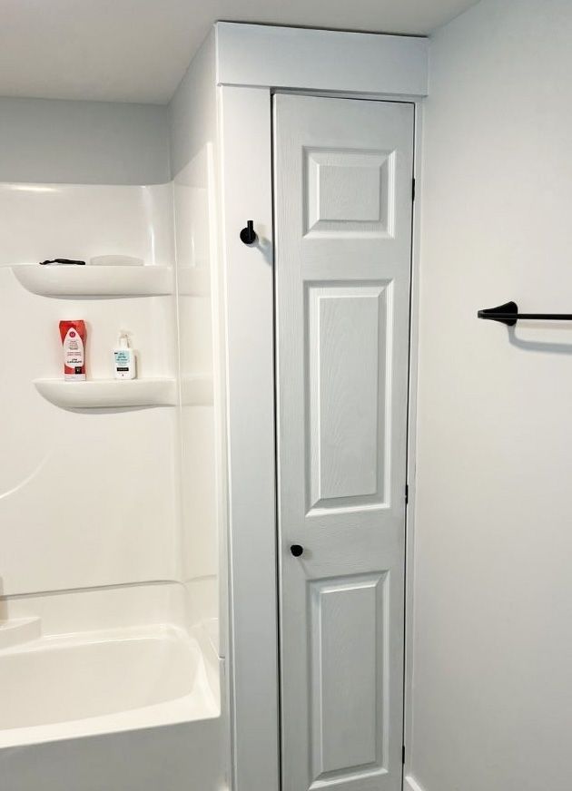 A white bathroom featuring a shower stall with built-in shelves, a narrow white door, and a black wall-mounted towel hook.