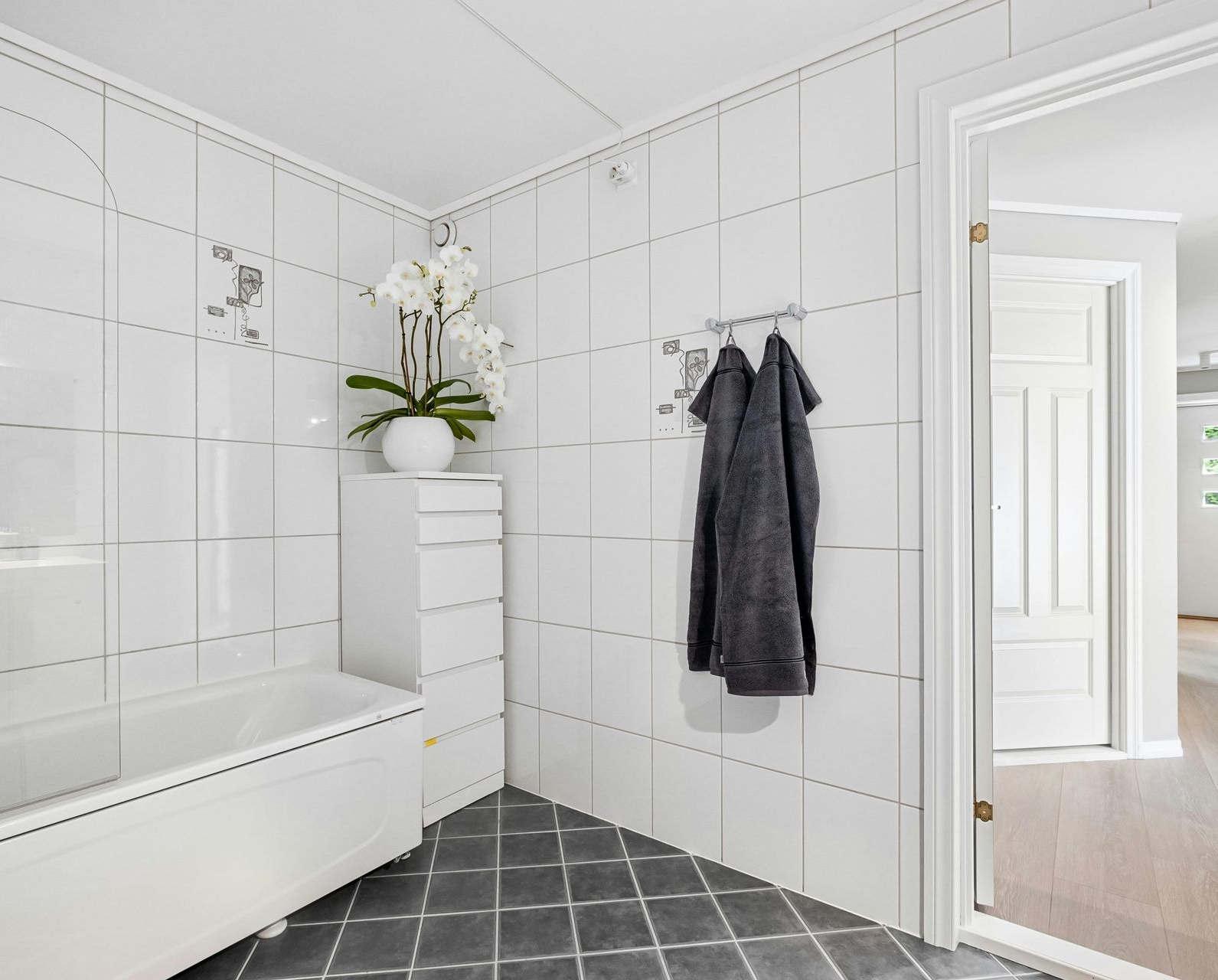 A bright bathroom with white-tiled walls, a white bathtub, a drawer unit with a white orchid, and a grey robe on a hook.