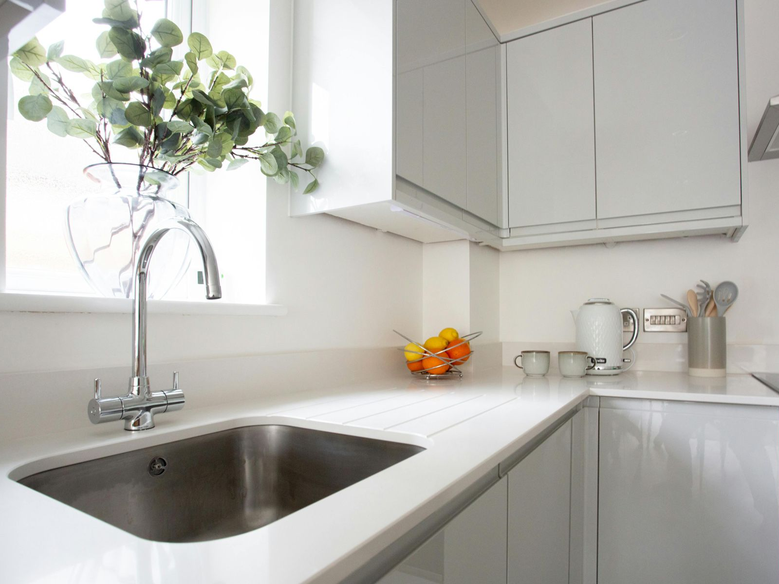 A bright, modern kitchen featuring a stainless steel sink, a chrome faucet, light grey cabinets, and a bowl of citrus.