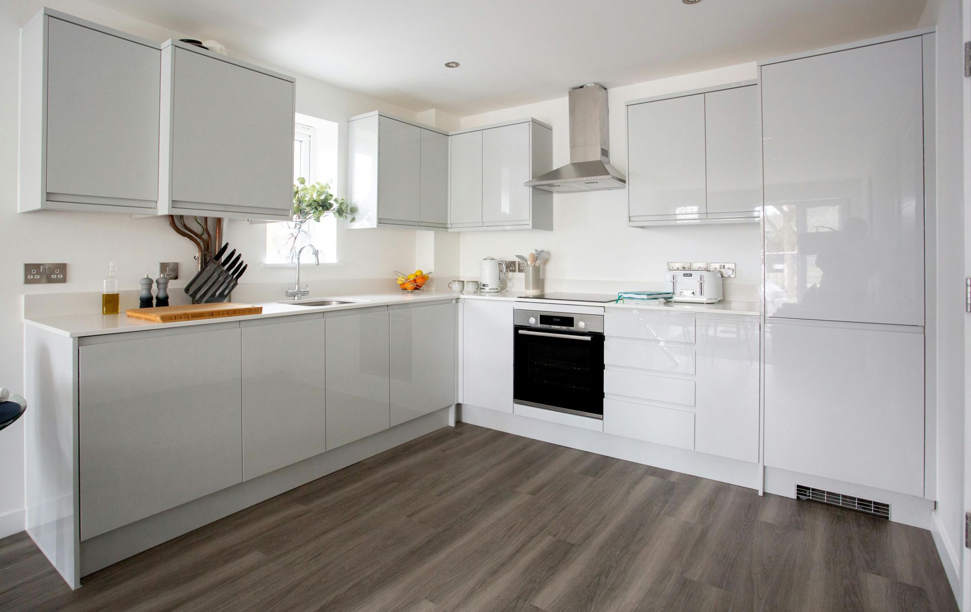 A modern kitchen with glossy white cabinets, stainless steel appliances, white countertops, and dark wood-look flooring.