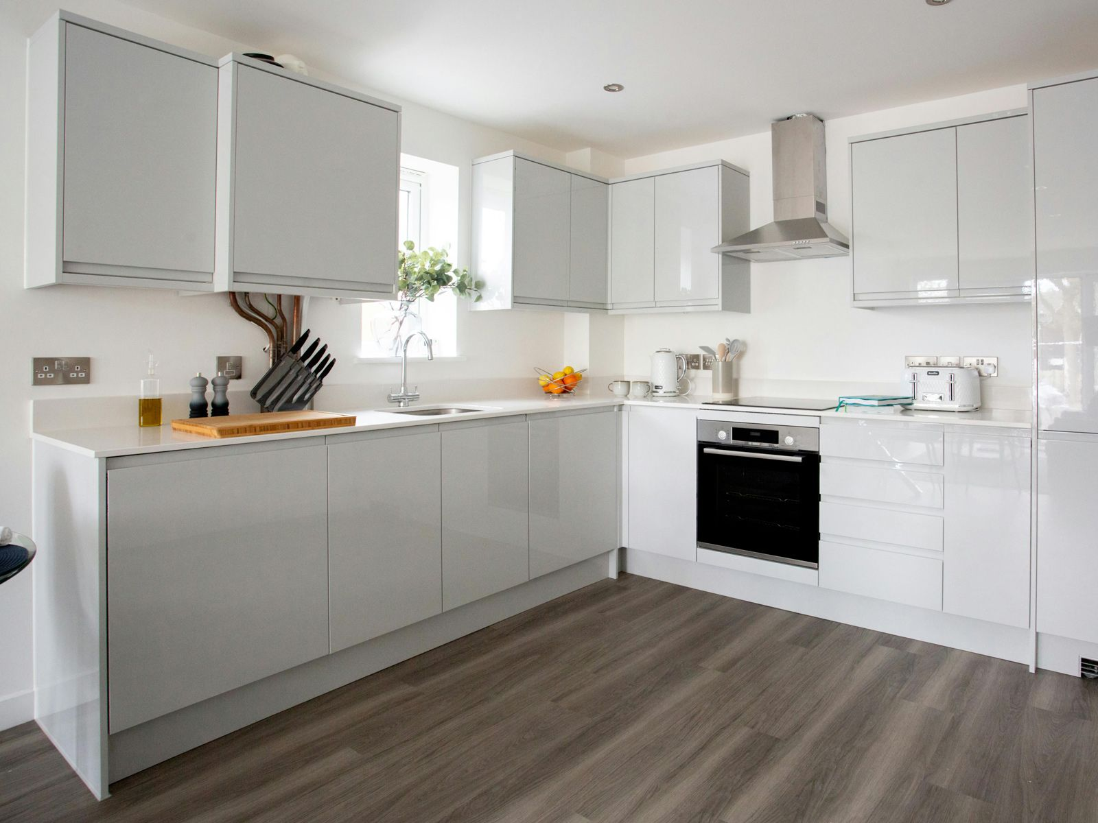 A modern kitchen with glossy white cabinetry, stainless steel appliances, white countertops, and dark wood-look flooring.