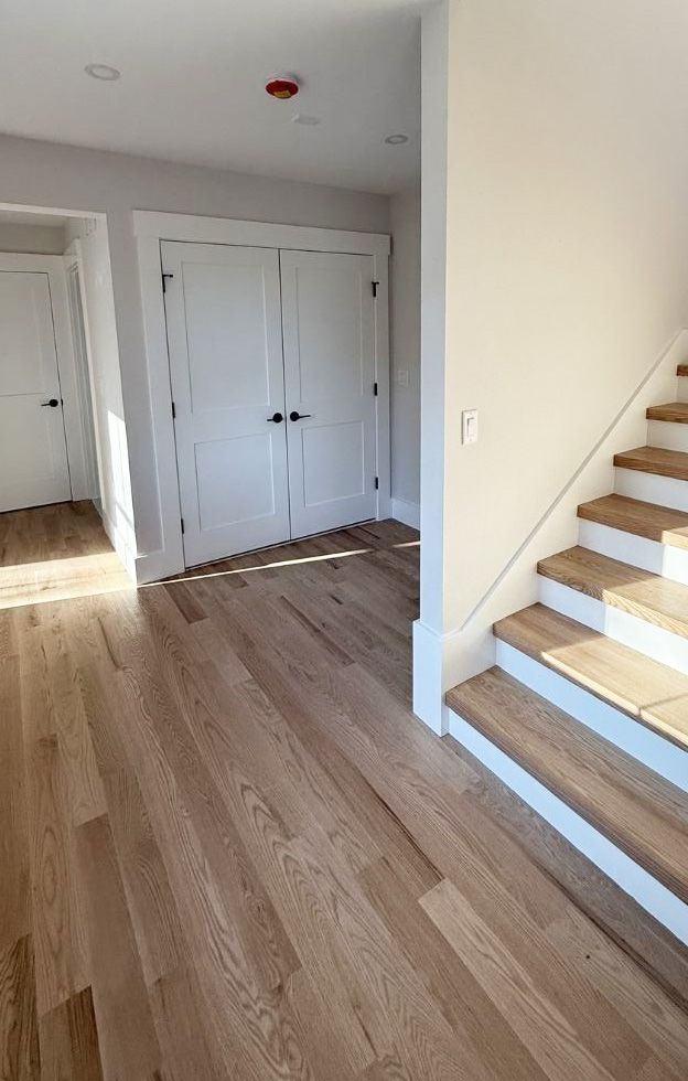 A bright interior hallway with light hardwood floors, white doors, and a staircase with wood treads and white risers.