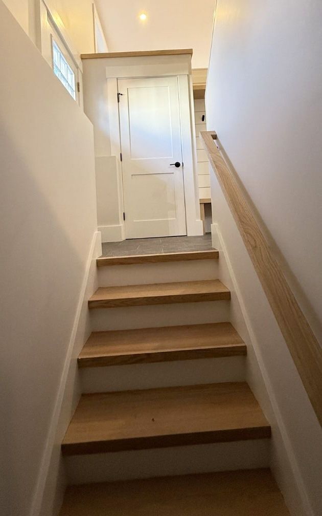 A staircase with white walls, wooden treads, and a wooden handrail leading up to a closed white door.