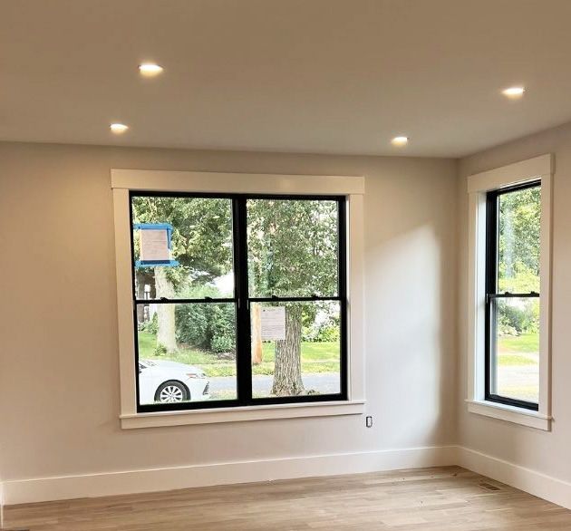 Interior view of a room with light beige walls, white trim, hardwood floors, recessed lighting, and two black-framed windows.