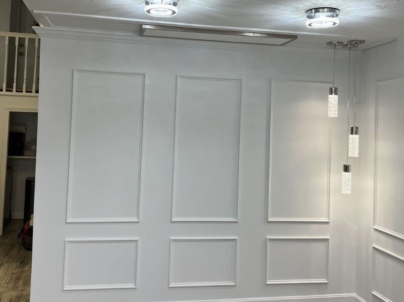 A white wall decorated with wainscoting panels, illuminated by two ceiling lights and three hanging pendant lamps.