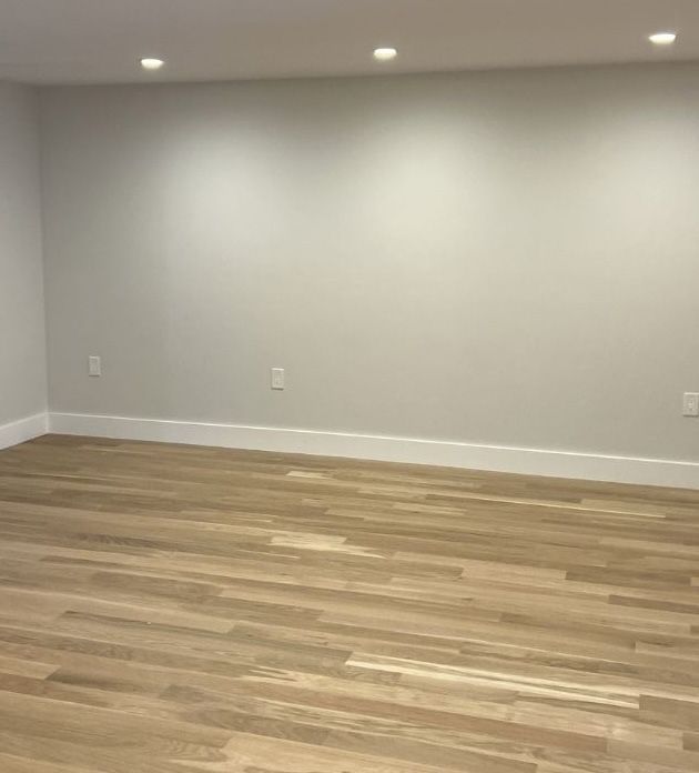 An empty room with light gray walls, white baseboards, light brown wood flooring, and recessed ceiling lighting.