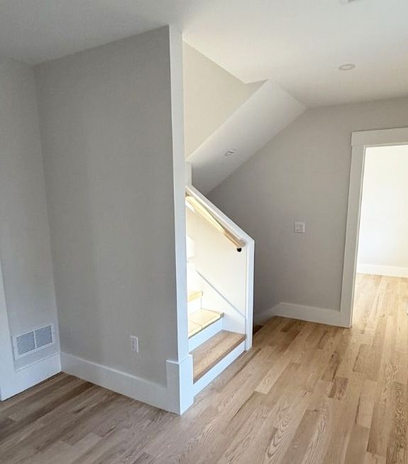 A staircase with light wood treads and a white railing, situated in a minimalist room with light gray walls and wood floors.