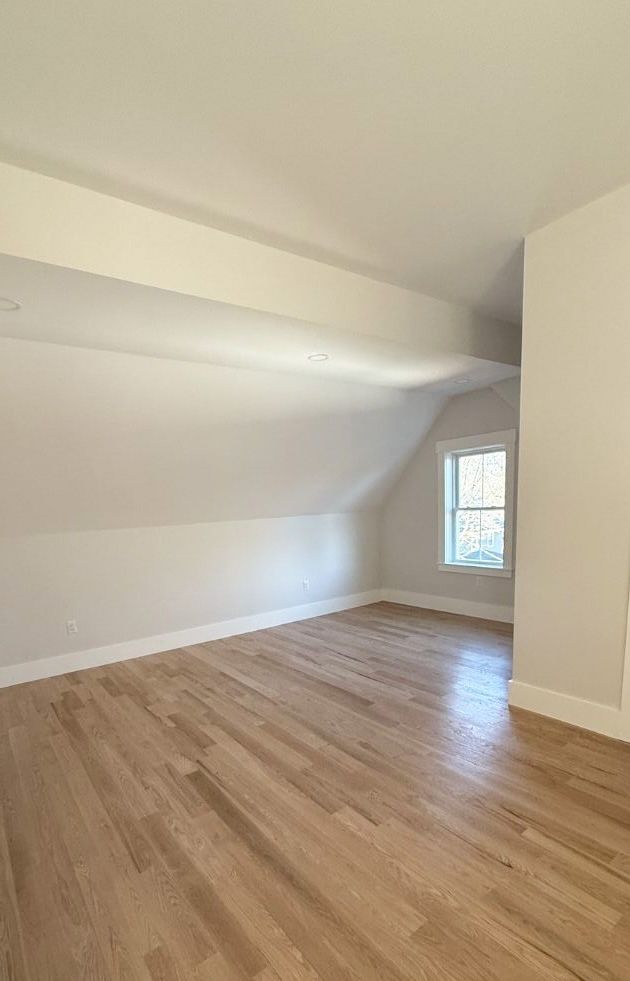 A bright, empty attic room with sloped ceilings, light-colored walls, light wood flooring, and a single window.