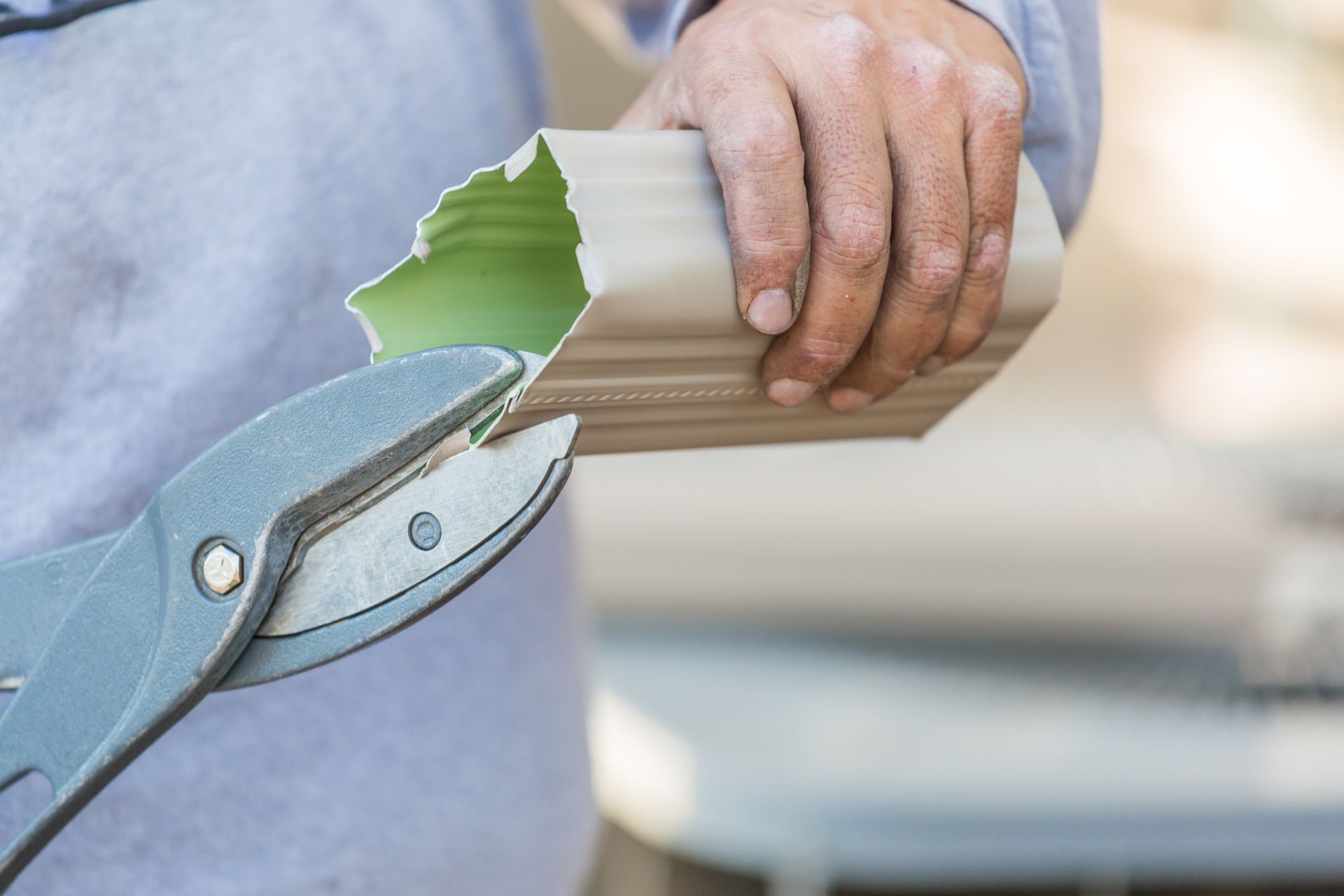 A person uses metal snips to trim a beige aluminum gutter downspout.