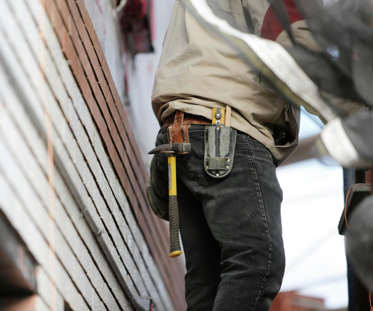 A person in work clothes with a hammer and tool holster on their belt, standing next to a structure under construction.