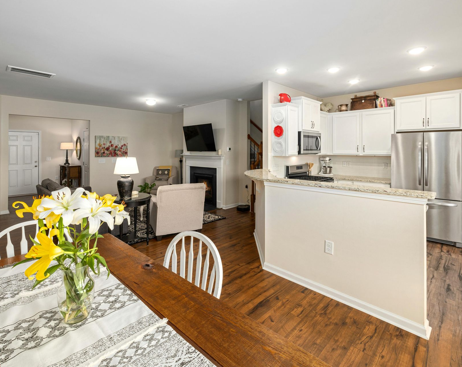 An open-concept living area with a foreground dining table, a kitchen with white cabinets, and a fireplace with a TV.