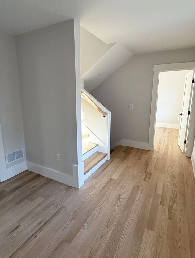 A view of a modern hallway with light hardwood floors, neutral gray walls, a white staircase, and an open doorway.