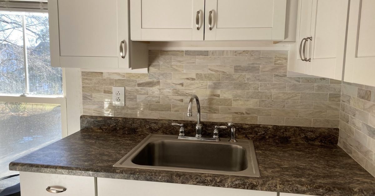 A kitchen sink with a chrome faucet set in a dark patterned countertop against a textured stone tile backsplash.