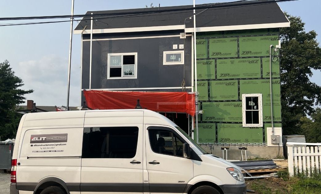 A white work van parked in front of a two-story house under construction, featuring partial siding and green sheathing.