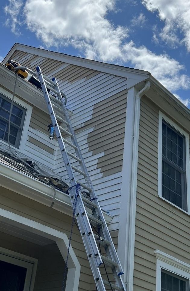 An extension ladder leans against the side of a two-story house with tan siding that is being painted white.