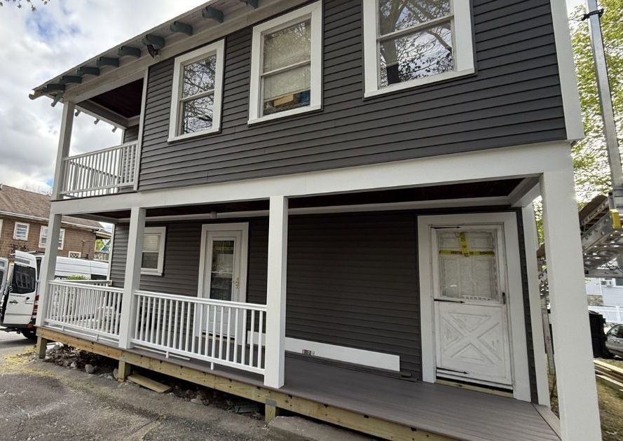 A two-story building with dark gray siding, featuring a white railed porch and two ground-level white doors.