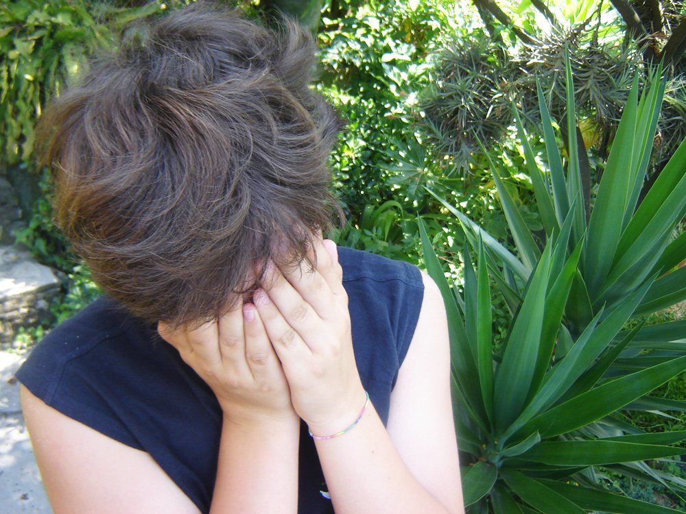 Person hiding face with hands in front of green plants; outdoors.