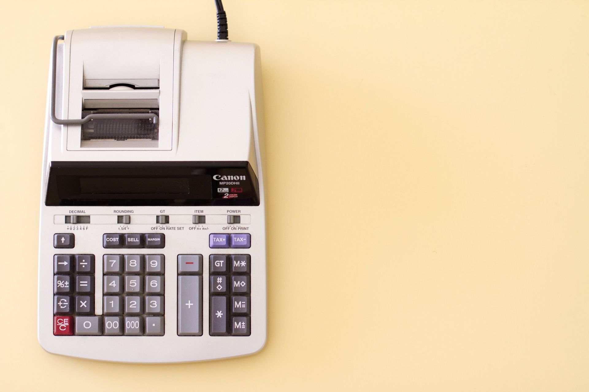 Adding machine on a light yellow background. The machine is light gray with black and colored buttons and paper roll.