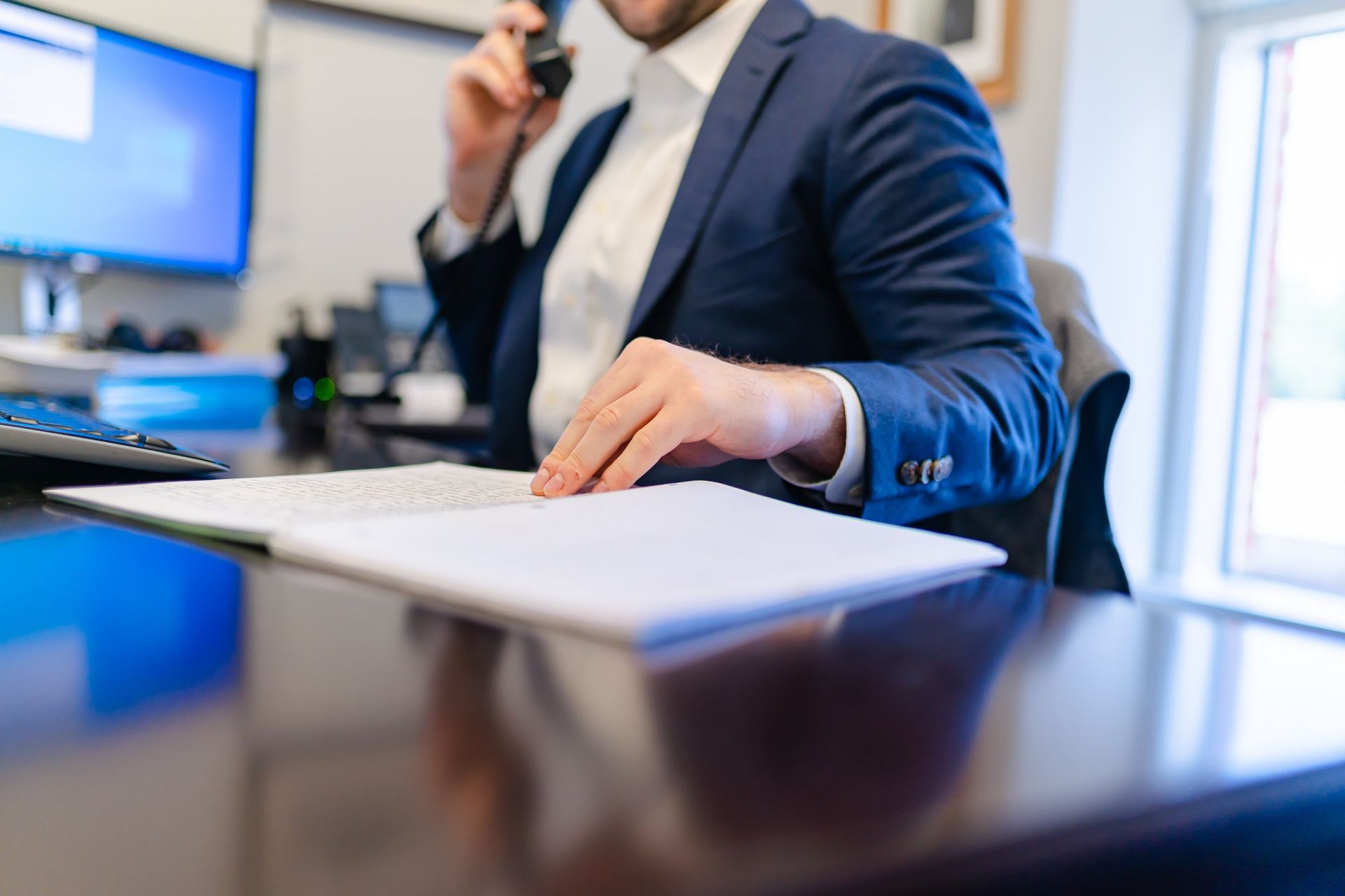 Man in suit on phone, reviewing paperwork at a desk in an office setting.
