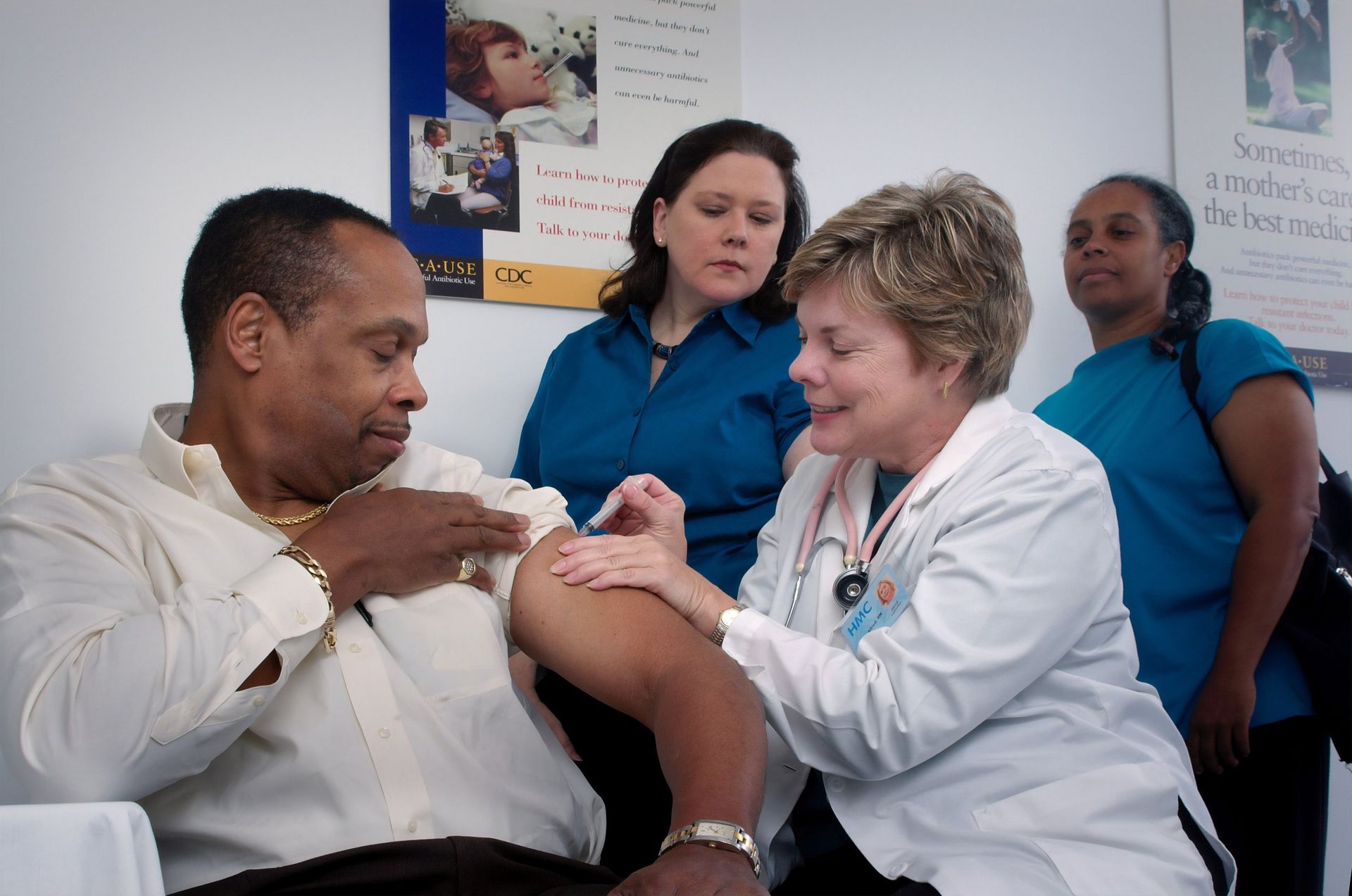 Doctor giving a Black man a shot in his arm; two other women watch in a clinic setting.