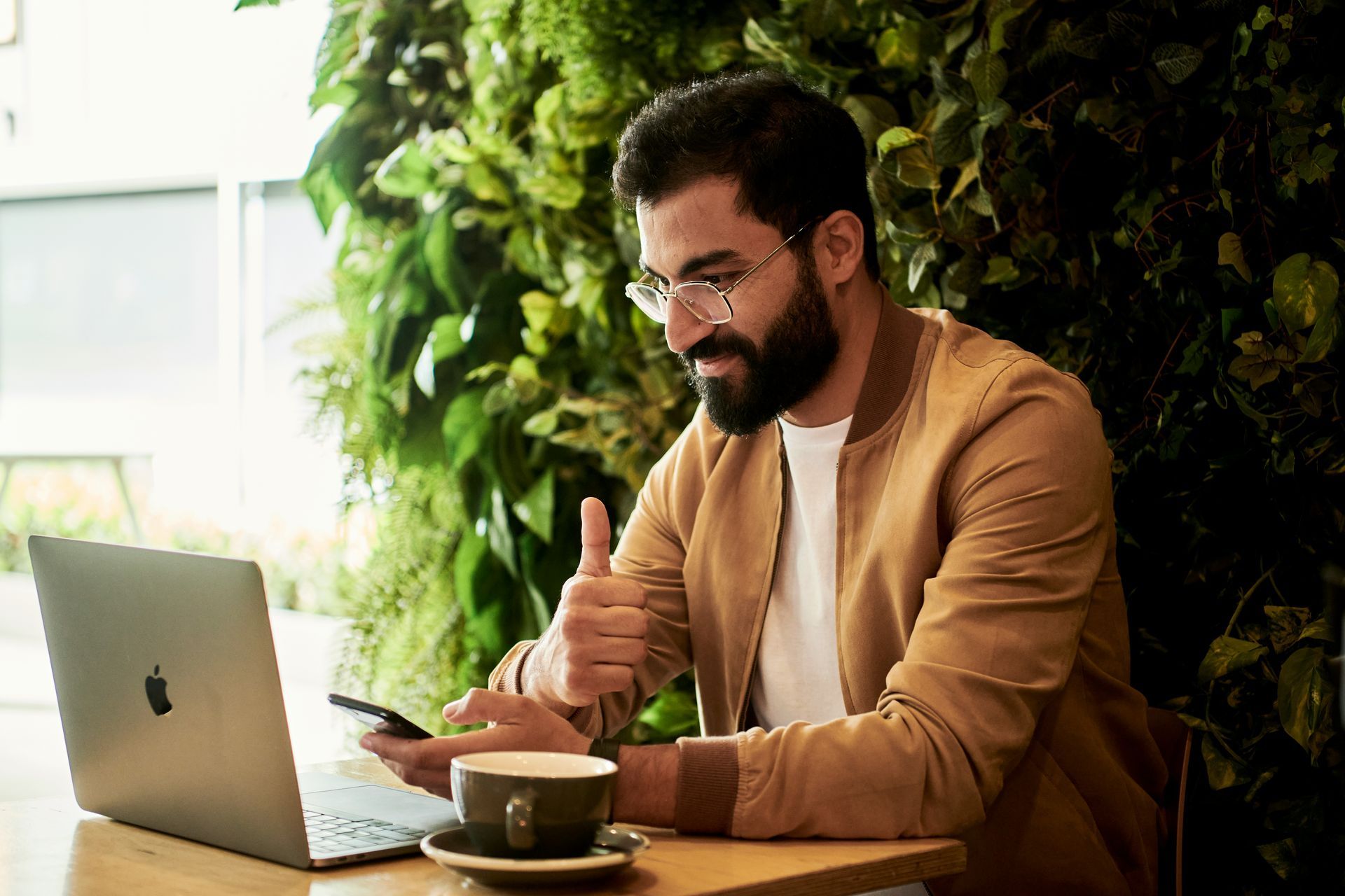 Man with glasses and beard giving a thumbs-up while working on laptop and phone at a cafe with a green wall.
