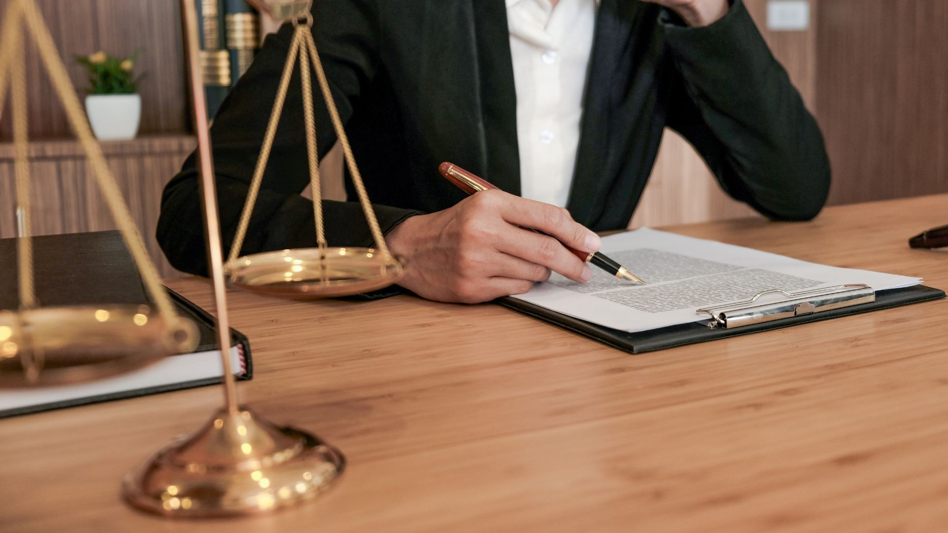 Lawyer in a dark suit writing on a document at a wooden desk with scales of justice.