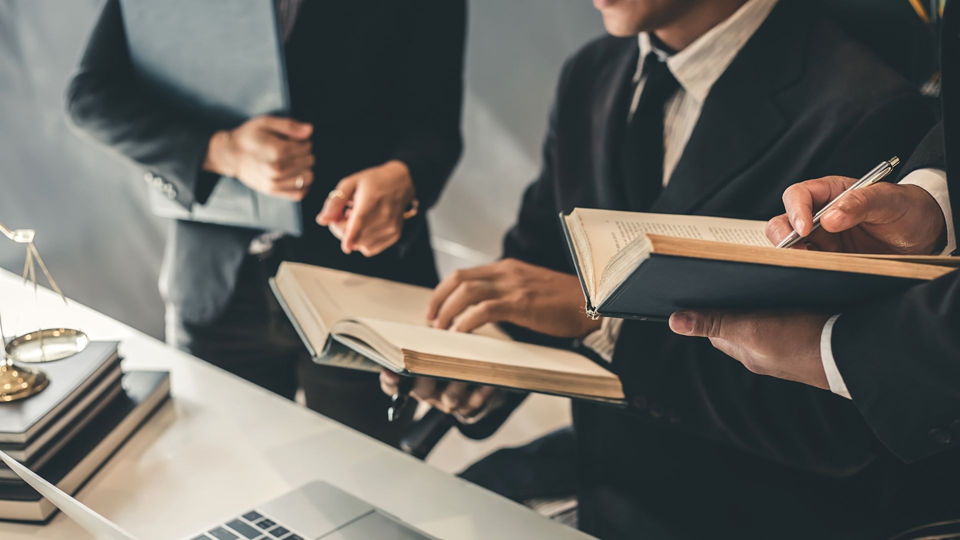 Lawyers in suits reviewing documents at a desk. Scales of justice and laptop visible.