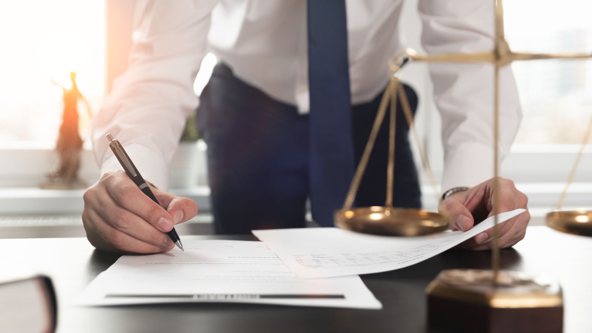 Lawyer signing documents next to scales of justice on a desk.