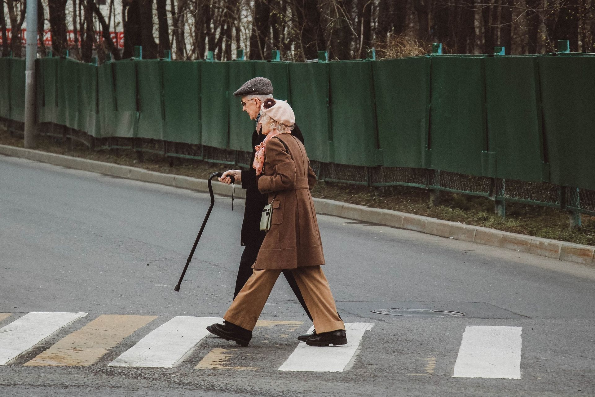 Elderly couple crossing a crosswalk, man with cane, brown coat, grey pants, green fence.
