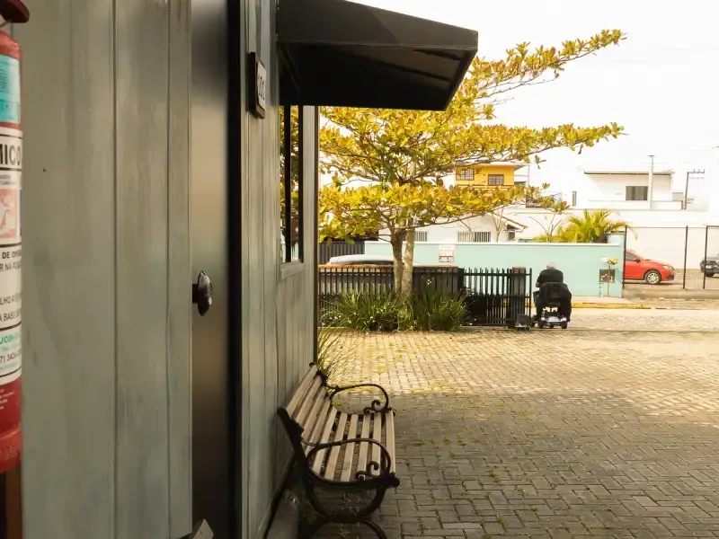 Vista exterior com um banco, um edifício, uma árvore e um carro estacionado numa área pavimentada com tijolos.