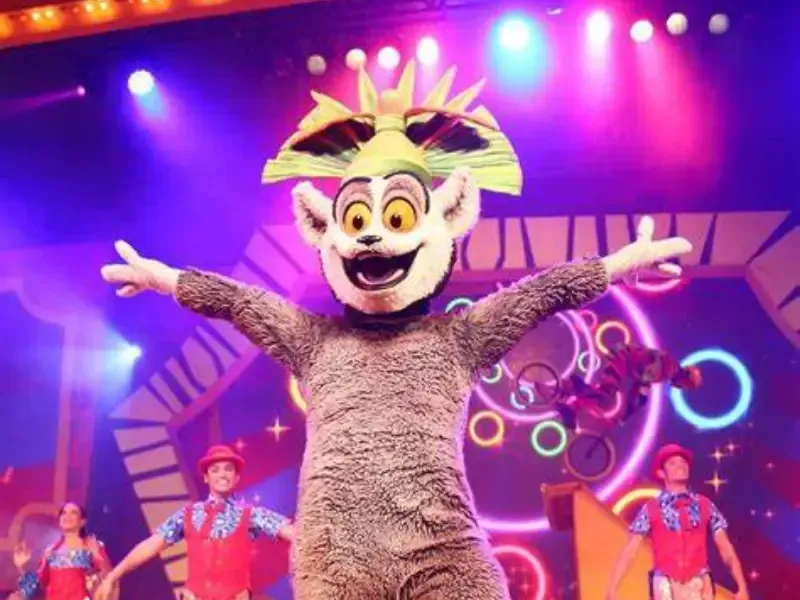 Personagem do Rei Julien em cena com os braços estendidos, rodeado por artistas, palco colorido.