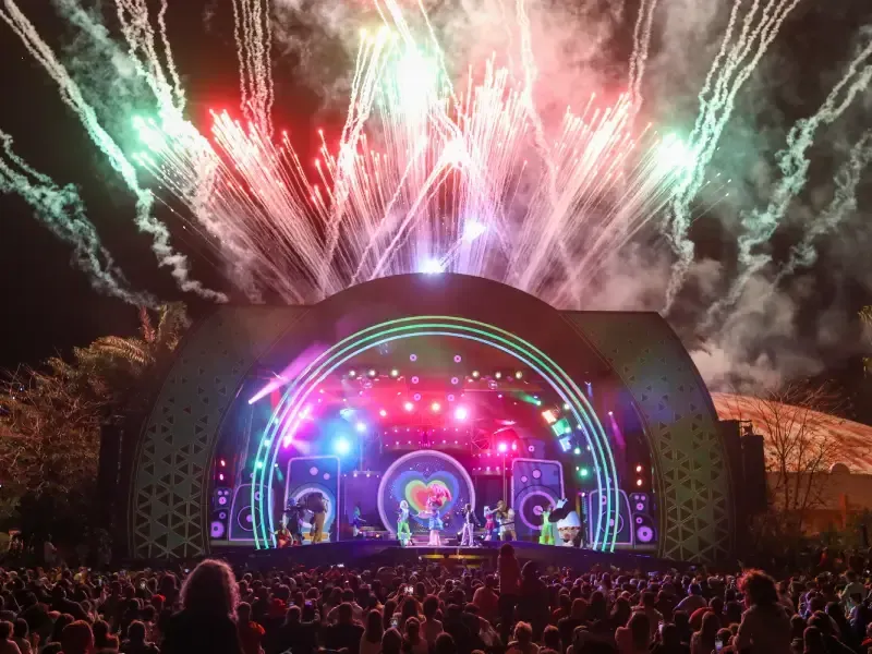 Palco de concertos com artistas, luzes coloridas e fogos de artifício sobre uma multidão.