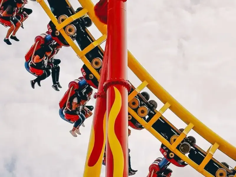 Carrinhos de montanha-russa com passageiros no ar, trilhos em vermelho e amarelo vibrantes contra um céu nublado.