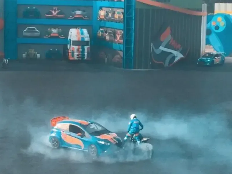 Carros de corrida azuis e laranjas derrapam, levantando fumaça, perto de uma pessoa, tendo como fundo peças de carros e logotipos.