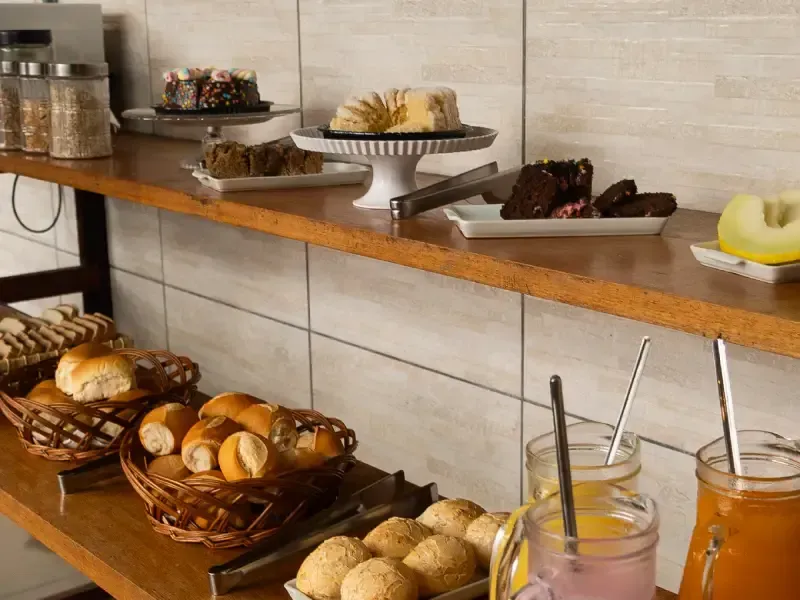 Buffet de café da manhã: pães, bolos, frutas e sucos em prateleiras de madeira, encostadas em uma parede de azulejos.