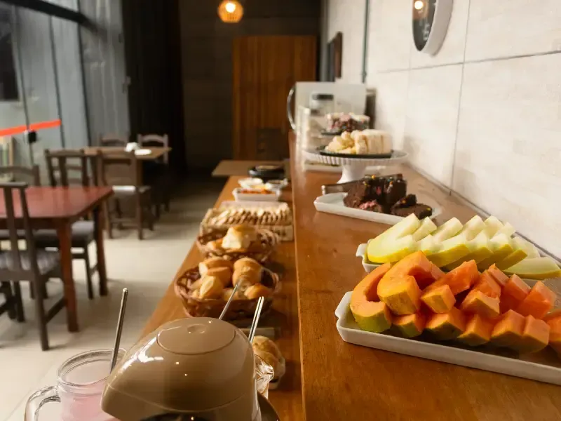 Buffet de café da manhã com doces, frutas e café sobre uma mesa de madeira. Mesas e cadeiras ao fundo.