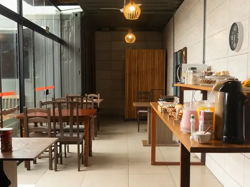 Interior de um pequeno café com mesas e cadeiras de madeira, balcão para refeições rápidas e luminárias pendentes.