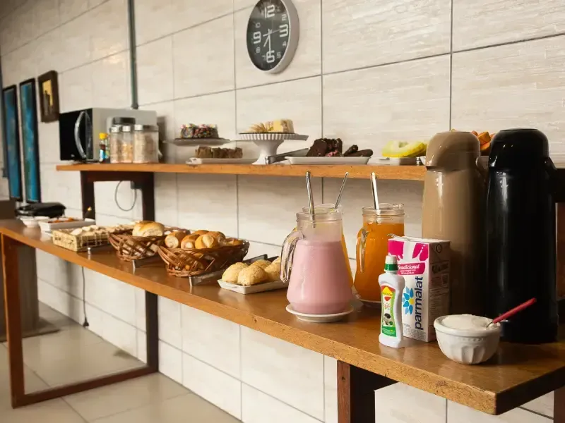 Buffet de café da manhã com pães, bebidas e micro-ondas em um longo balcão de madeira.