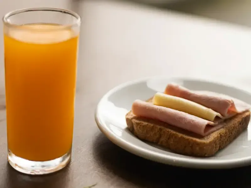 Um copo de suco de laranja ao lado de um prato com um sanduíche de presunto e queijo em uma torrada.