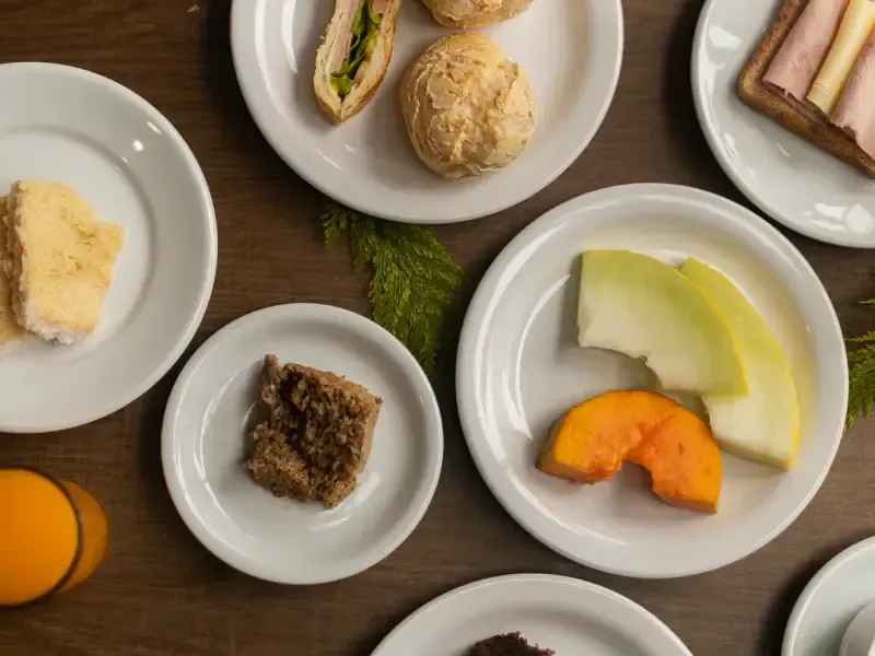 Vista aérea de diversos alimentos para o café da manhã em pratos brancos: torradas, frutas, doces e suco de laranja sobre uma mesa de madeira.