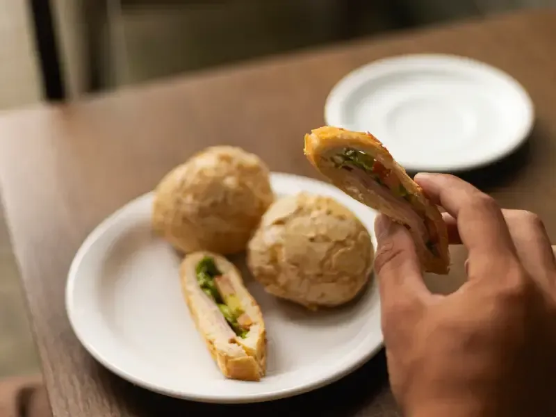 Pessoa segurando um sanduíche, com outros itens assados ​​redondos e um prato vazio sobre uma mesa.