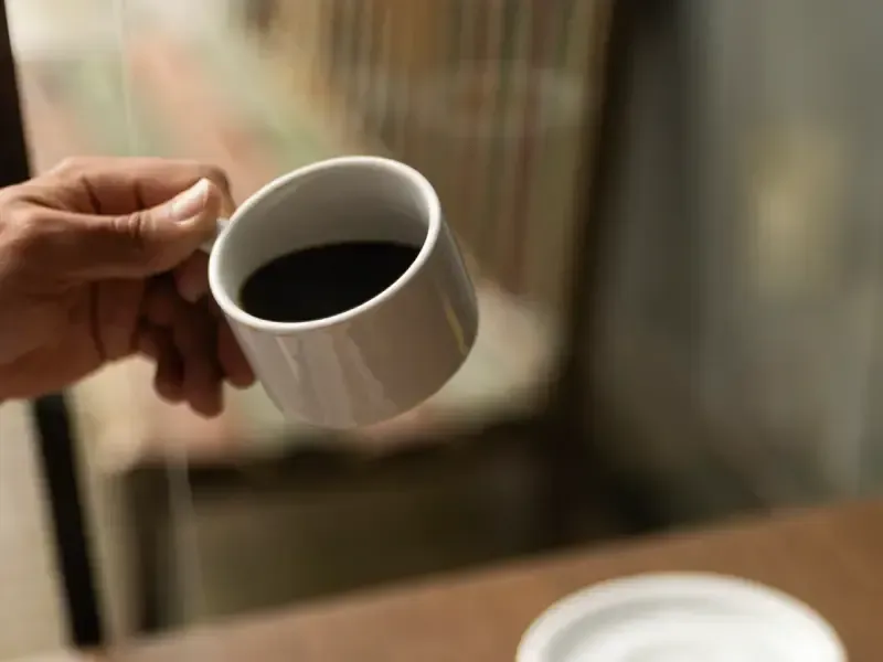 Mão segurando uma caneca branca com um líquido escuro, possivelmente café, em ambiente interno.