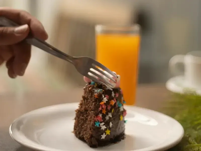 Uma garfada de bolo de chocolate com granulado, suco de laranja e uma mão.