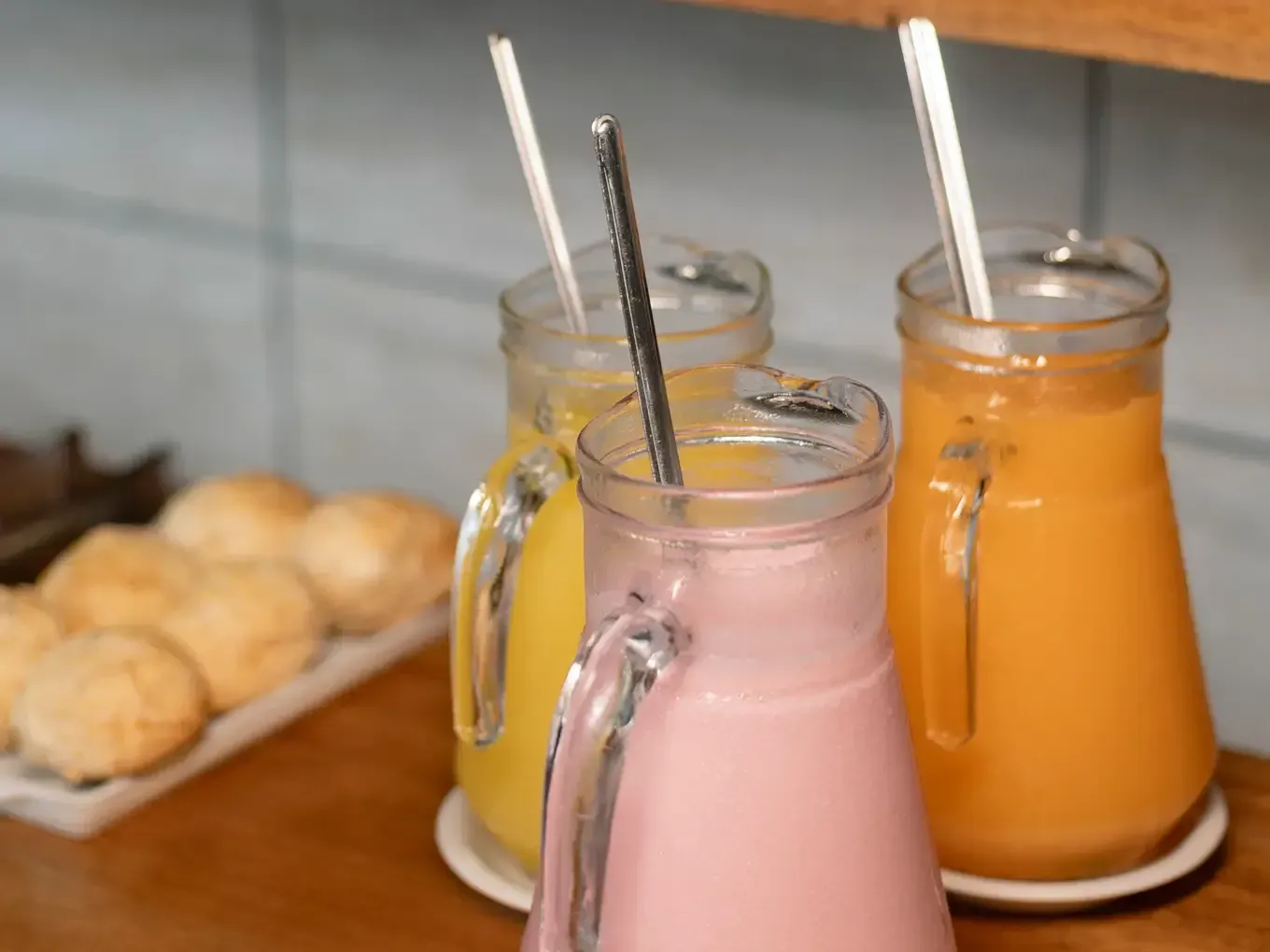 Três smoothies coloridos em potes de vidro com mexedores e pãezinhos em uma bandeja.