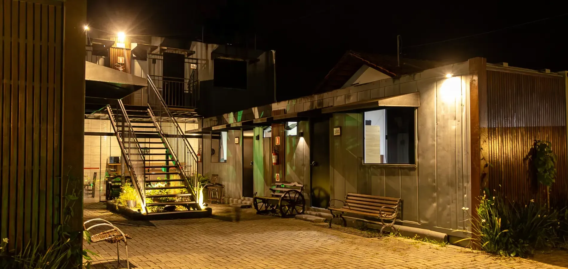 Exterior de um prédio à noite, com escadas, uma pequena mesa e um banco. Iluminado por luzes no teto.