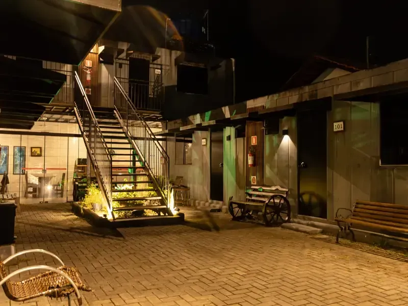 Pátio à noite com escadaria metálica, pavimentação em tijolo e edifícios em estilo industrial.