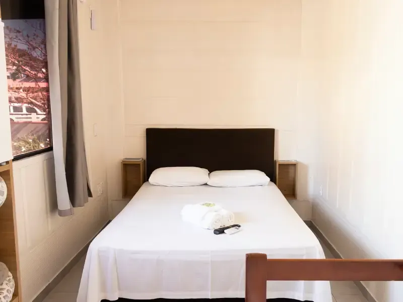 Quarto com cama de lençóis brancos, cabeceira marrom, mesas de cabeceira e janela com cortina cinza.