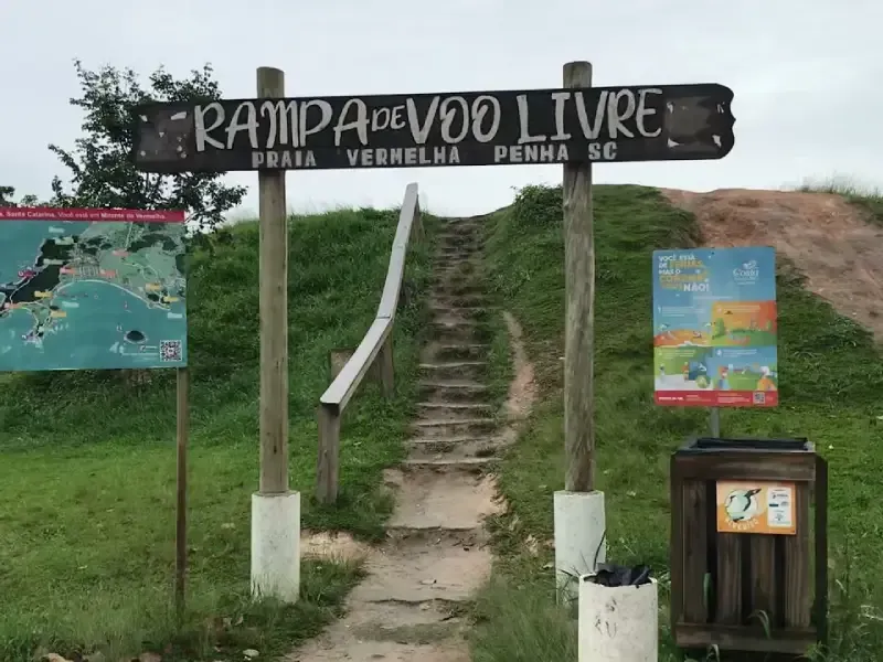 Placa de madeira indicando uma rampa de voo livre na Praia Vermelha, Penha, SC, com escadas que levam a uma colina gramada.