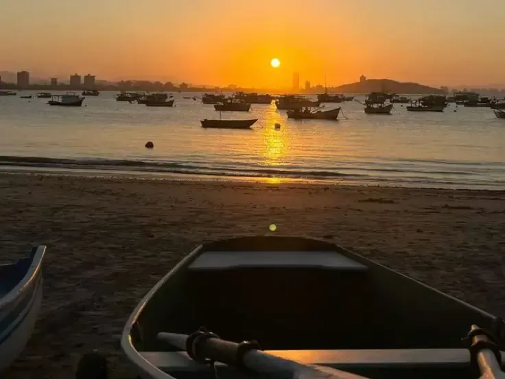 Pôr do sol sobre uma baía tranquila com barcos de pesca; um pequeno barco a remo está atracado na praia.