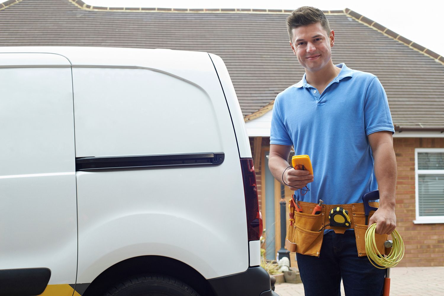 Electrician with a tool in his hands, wearing a tool belt beside his van with a house in the background. Electrician with a tool in his hands, wearing a tool belt beside his van with a house in the background.